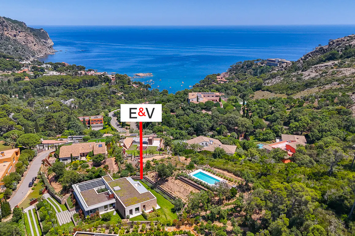 Aerial view of a modern villa with a pool, surrounded by lush greenery and hills overlooking the blue Mediterranean Sea.