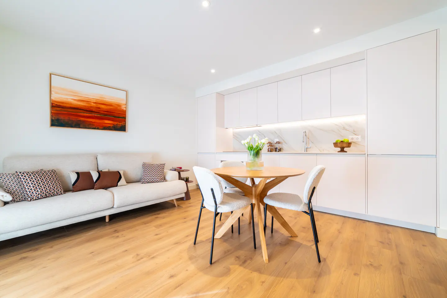 Bright, modern apartment interior with a beige sofa, round wood table, and white kitchen cabinets. An orange abstract painting hangs on the wall.