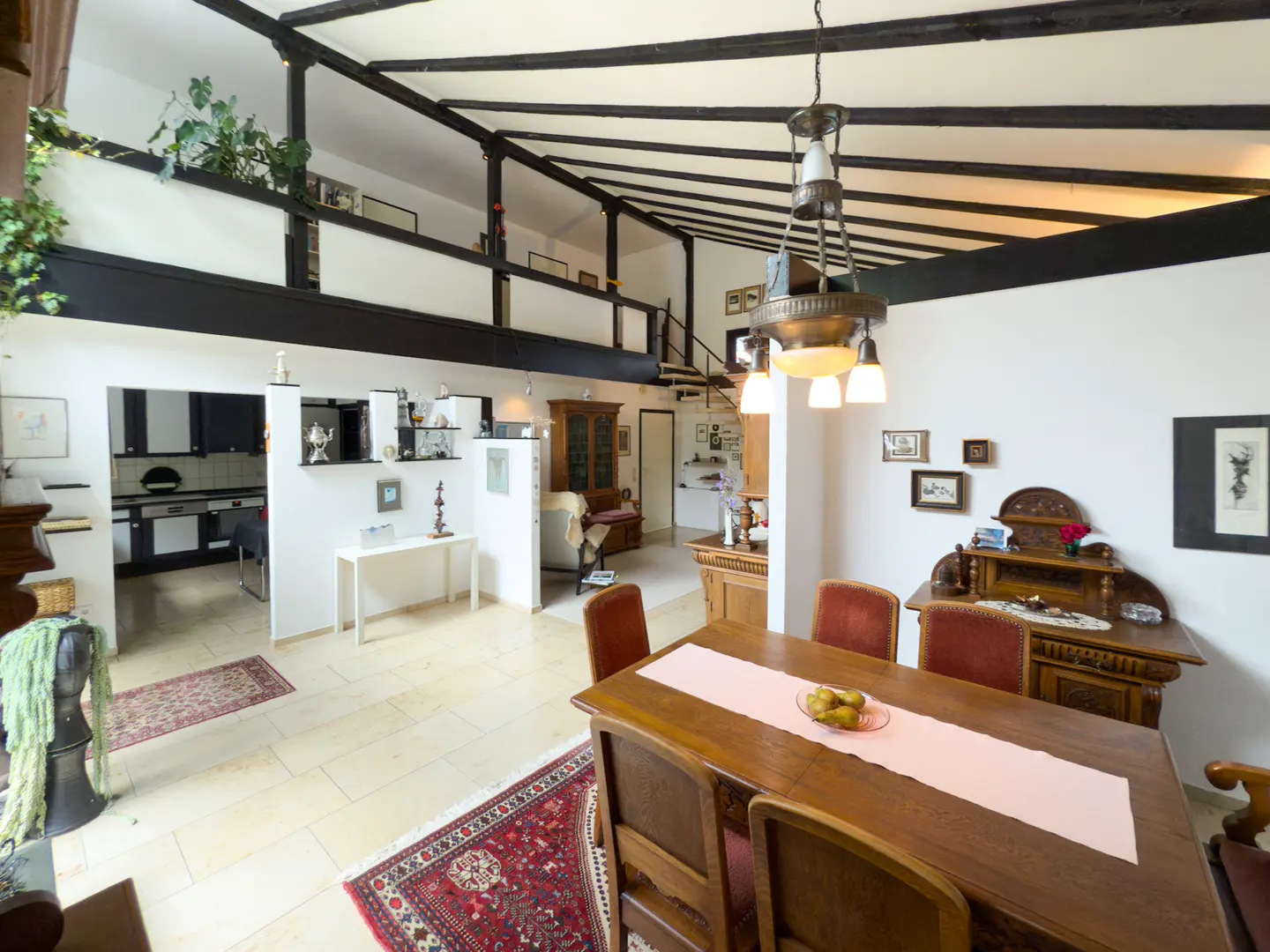 A bright, open dining room with a wooden table, red chairs, and a vintage chandelier. Black beams support a loft with plants.