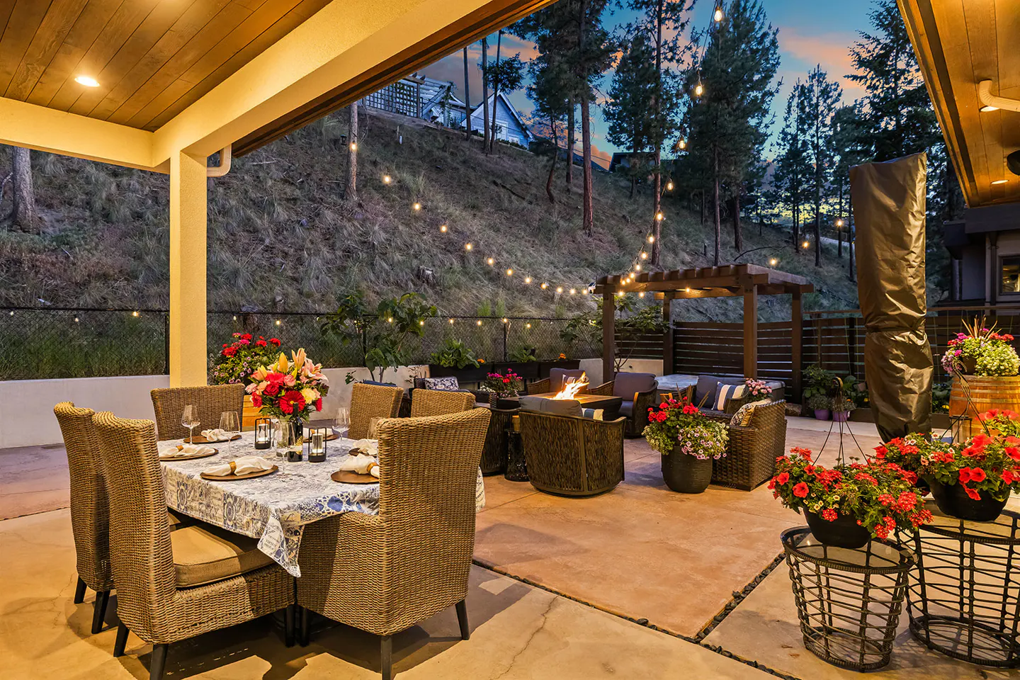 Outdoor patio with dining table, fire pit, and lounge area. String lights hang above, with a hillside and trees in the background.