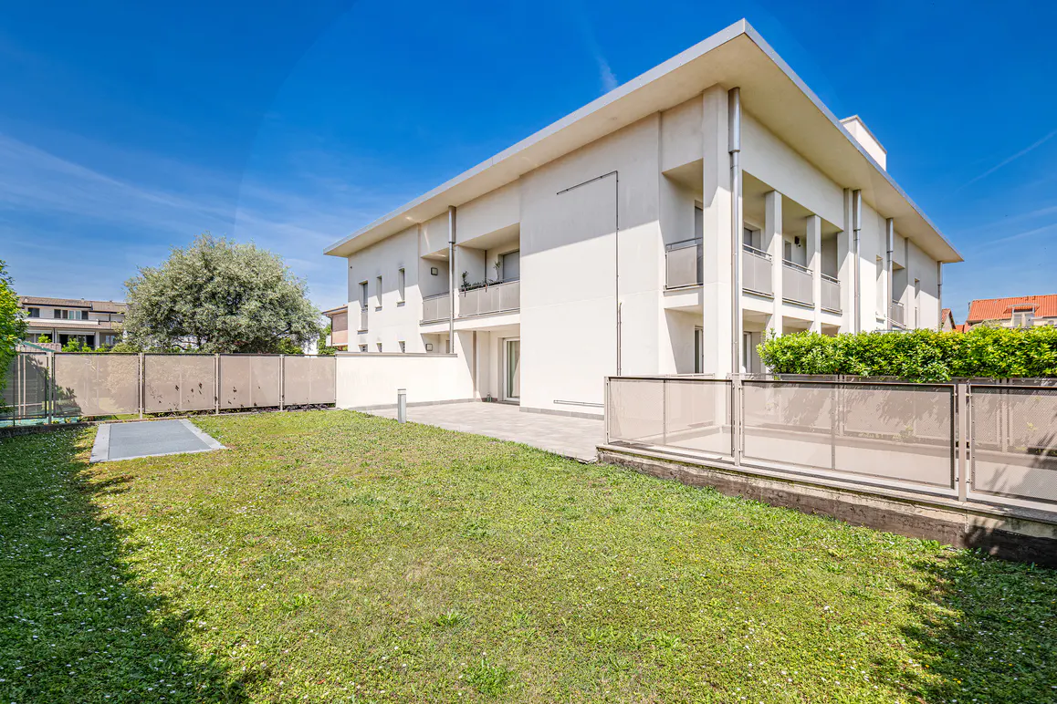 Modern white building with balconies, viewed from a grassy yard under a blue sky. A fence and trees are in the background.