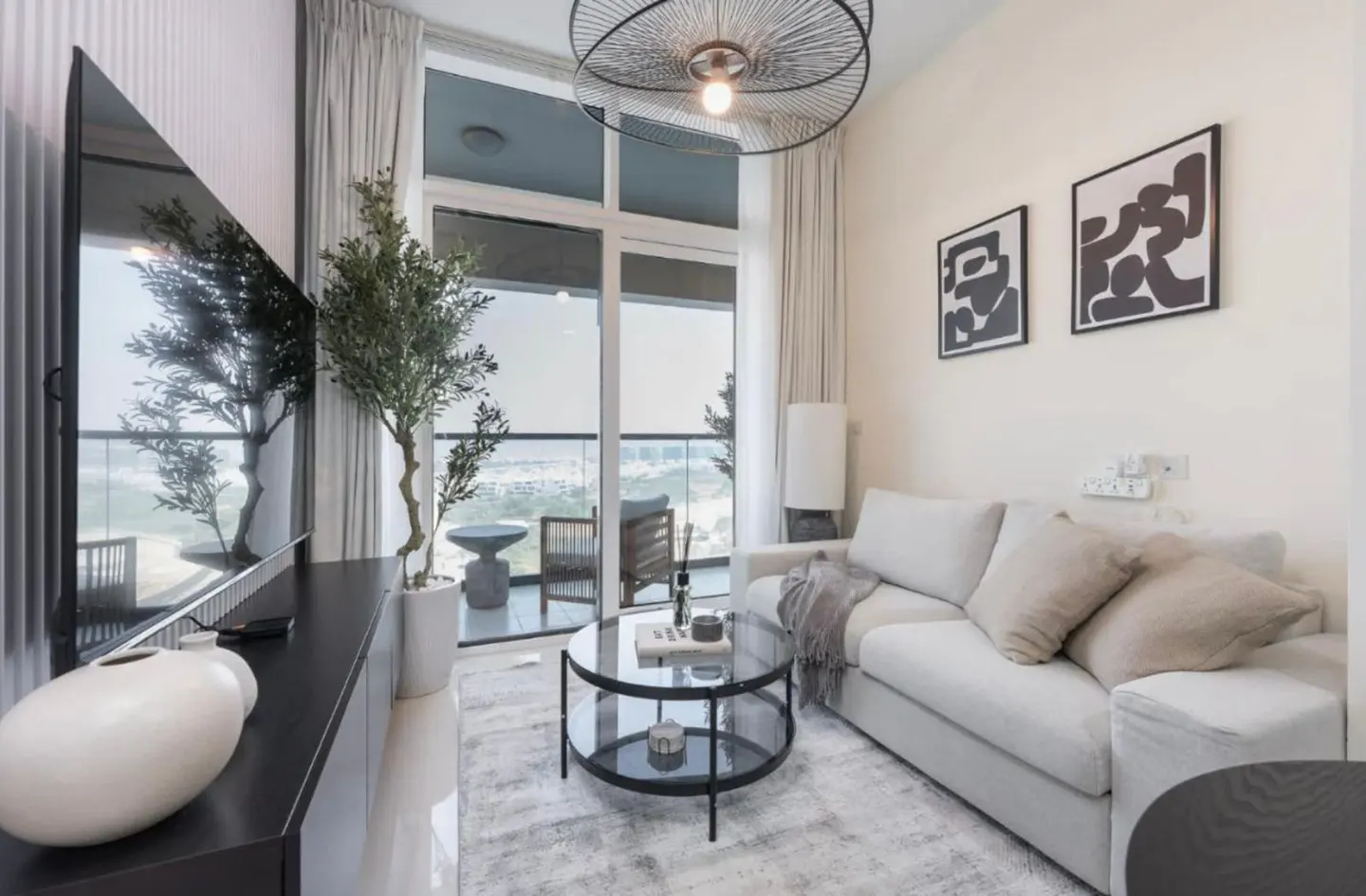 Living room with a white sofa, glass coffee table, and balcony view. Abstract art hangs on the wall. A modern chandelier illuminates the space.