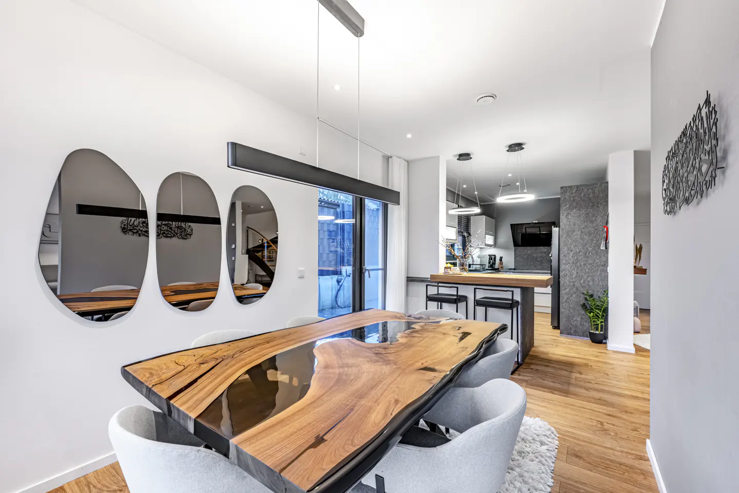 Bright dining area with a live-edge wood table, gray chairs, and modern lighting. Kitchen visible in the background.