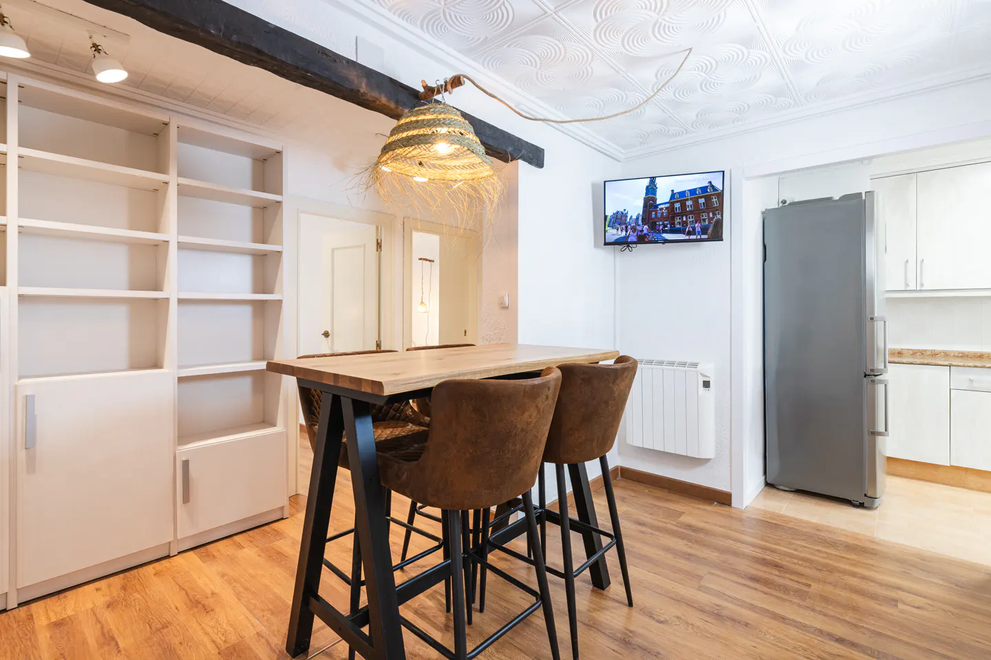 A kitchen with a wooden table, brown chairs, a straw lamp, and a TV on the wall.