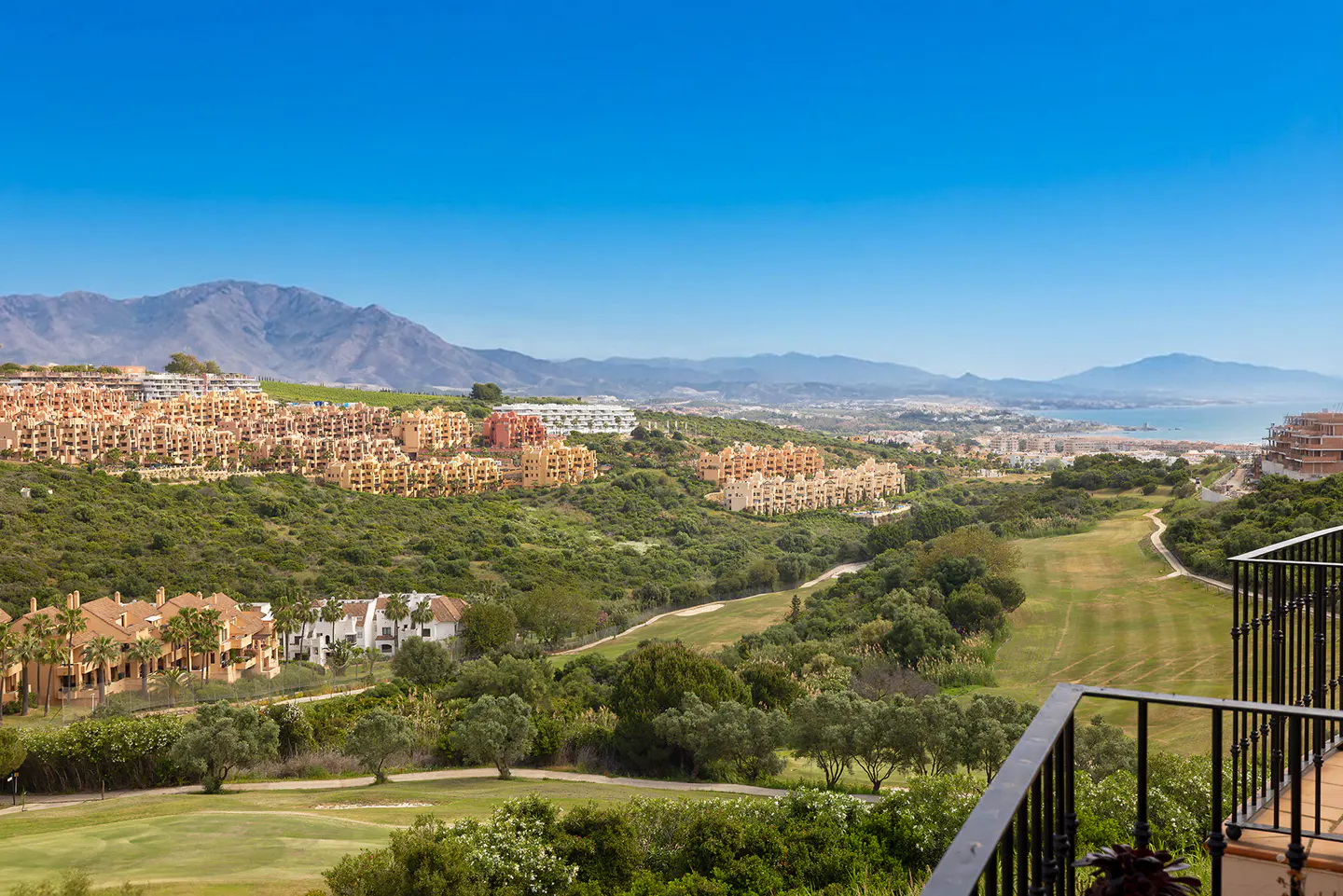 View from a balcony overlooking a golf course, mountains, and tan buildings under a clear blue sky.