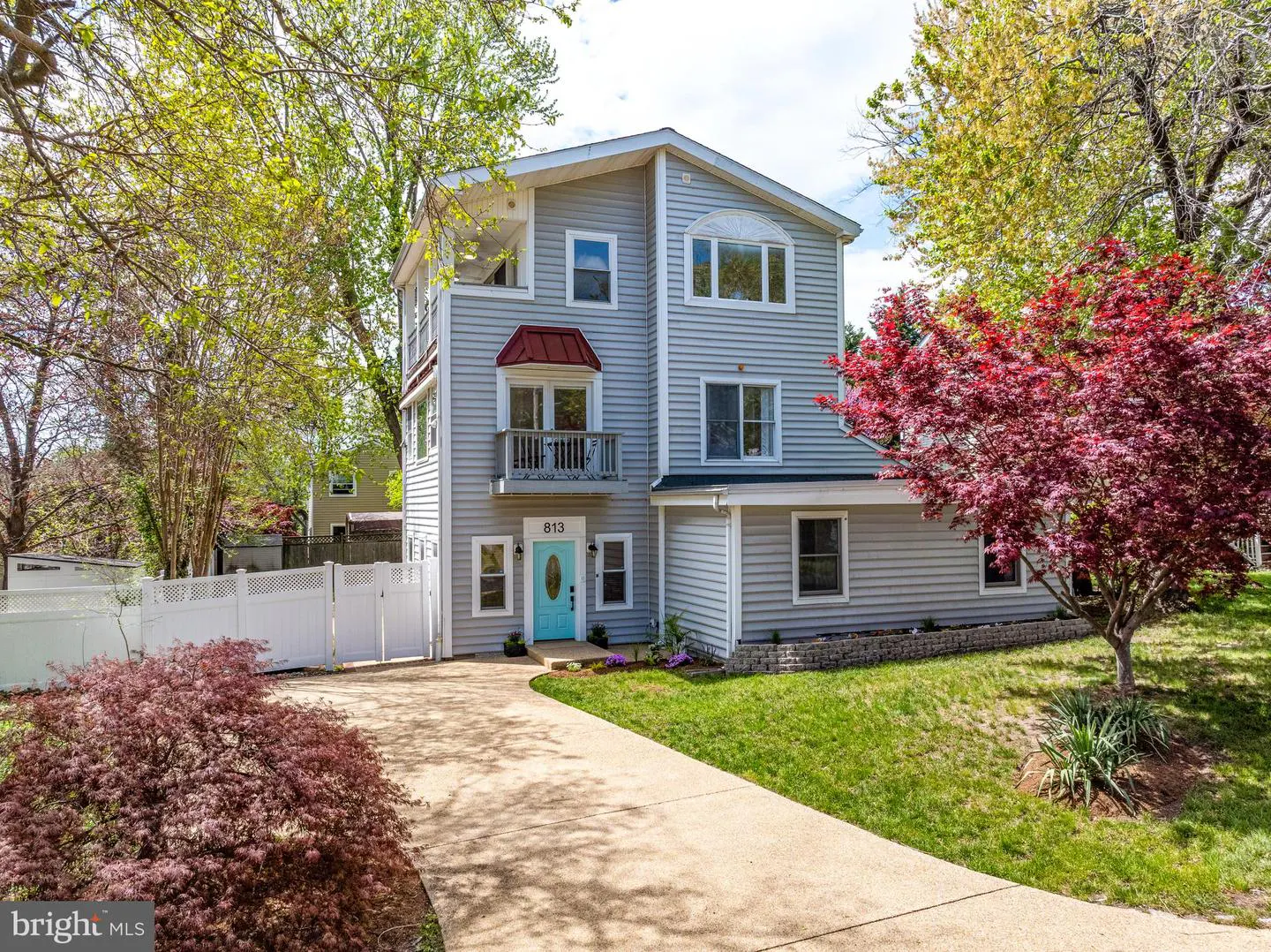 Two-story gray house with a blue door and red awning. A concrete driveway leads to the house, surrounded by green grass and trees.