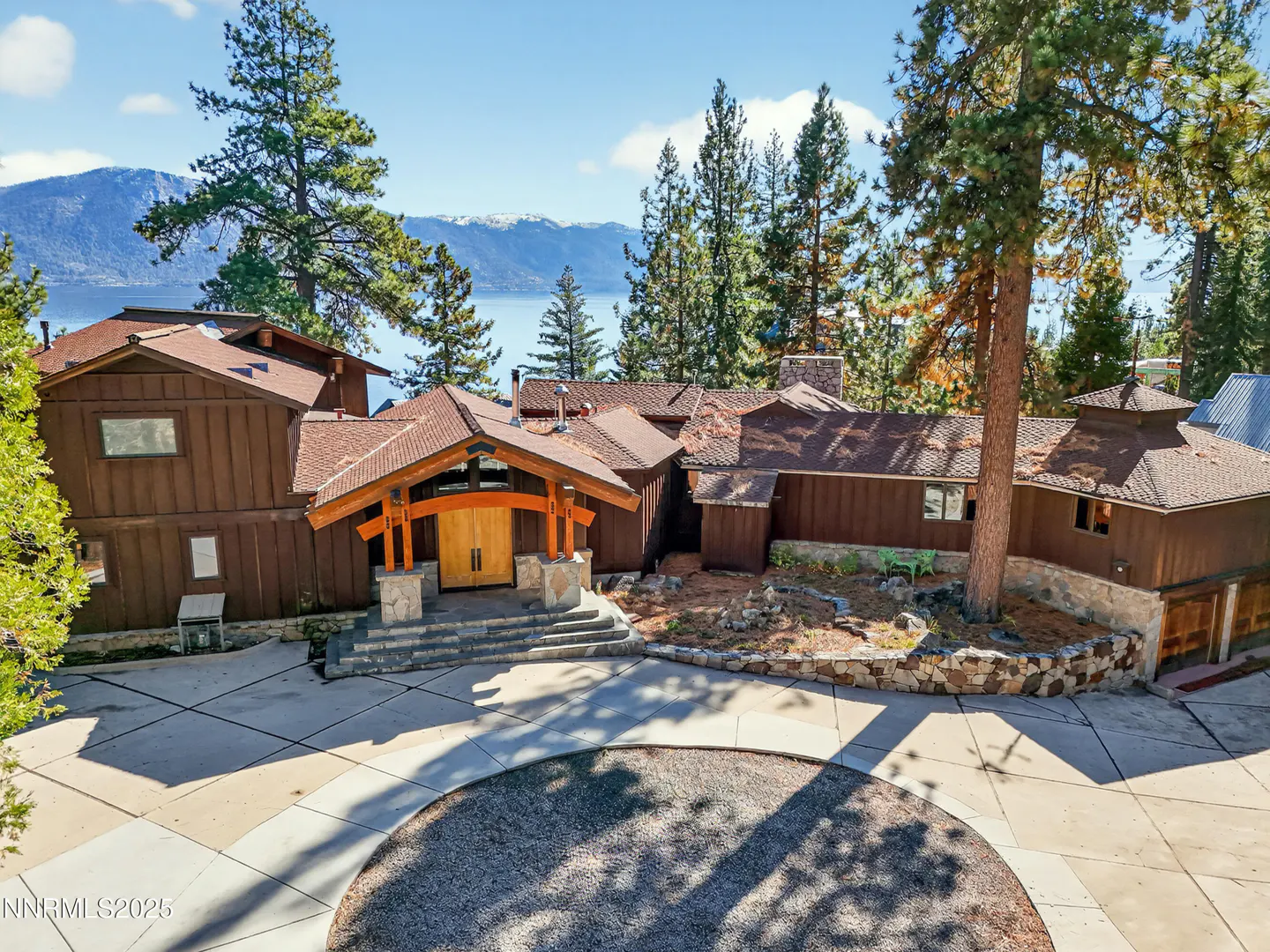Aerial view of a brown house with a circular driveway, trees, and a lake in the background.