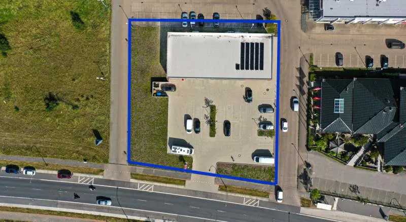 Aerial view of a commercial building with a white roof, solar panels, and a parking lot with several cars. A blue line marks the property's boundary.