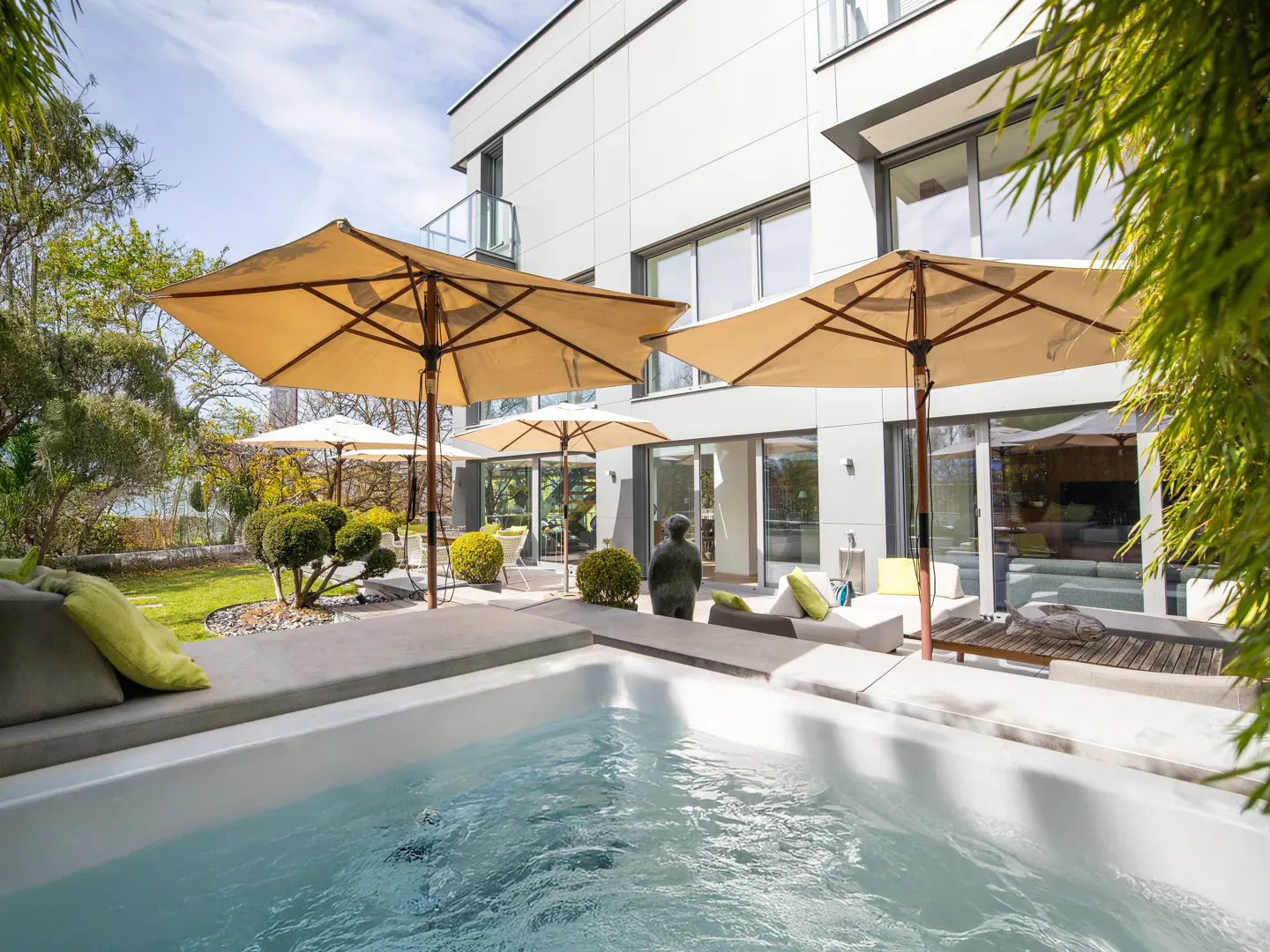 Modern backyard with a white hot tub, tan umbrellas, and lounge seating. A statue sits near the sliding glass doors of the gray house.