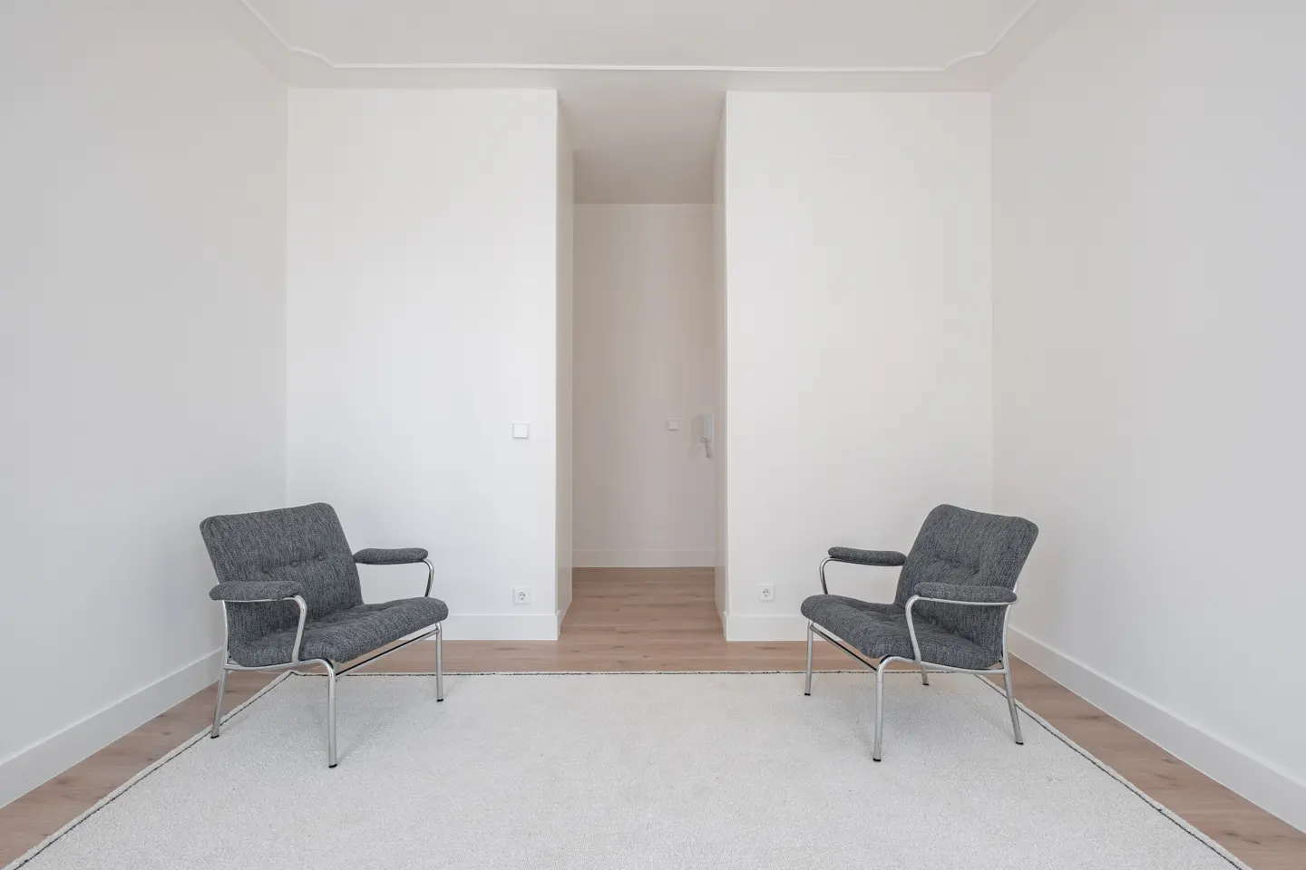 Two grey armchairs with metal frames face each other on a white rug in a minimalist room with white walls and wood floors.