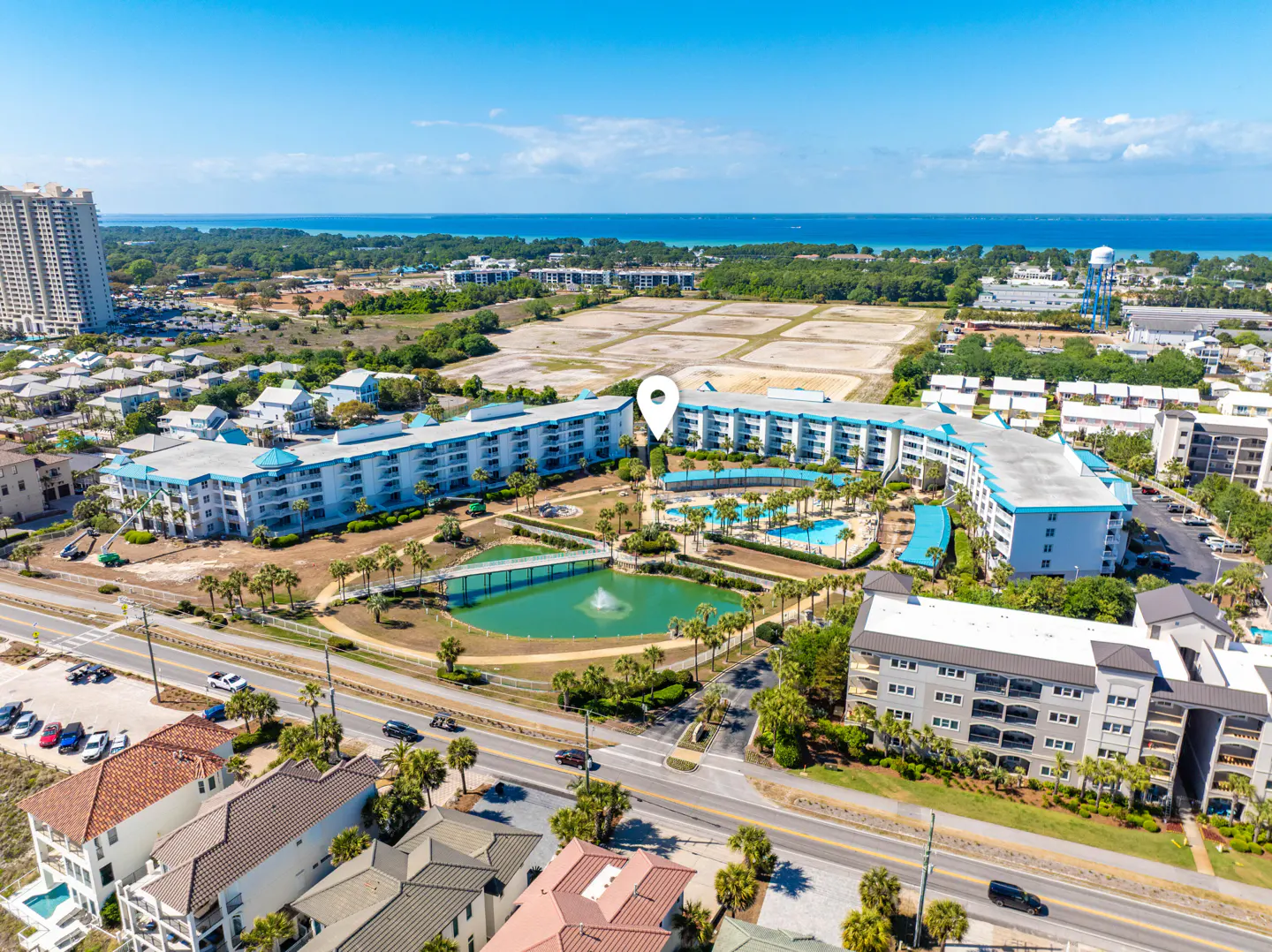 Aerial view of a white condo complex with a pool, pond, and palm trees near a beach with blue water.