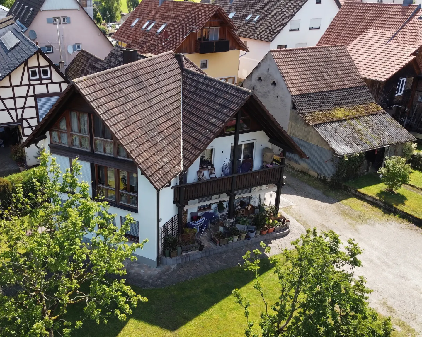 Two-story white house with a brown tiled roof and balcony, surrounded by green trees and other houses.