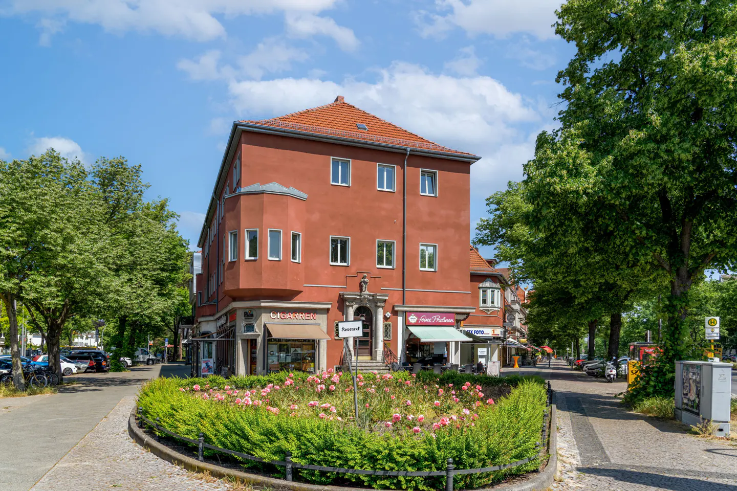 A three-story red building with a red tile roof, surrounded by trees and a flower garden. Shops are on the ground floor.