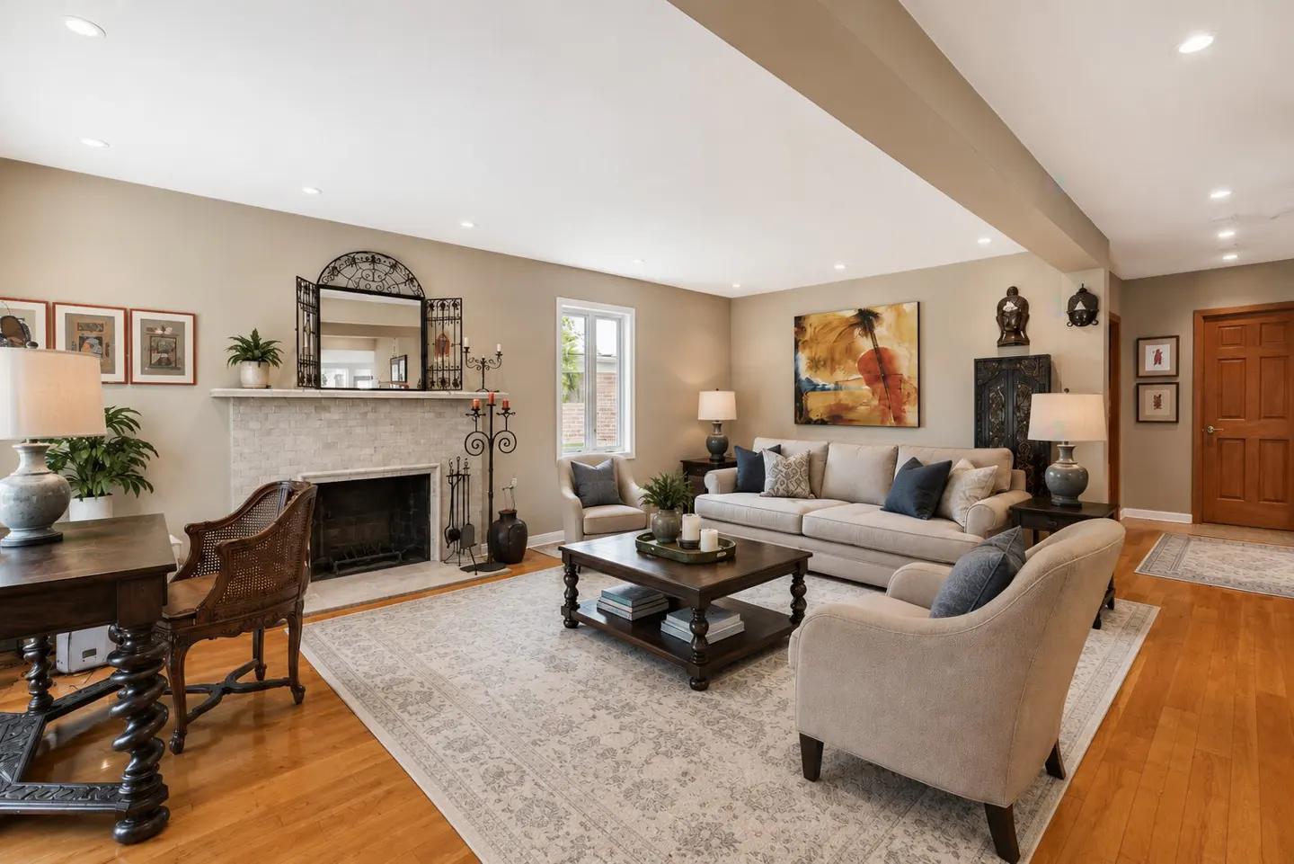 Spacious living room with a stone fireplace, beige sofa, armchairs, wooden desk, and hardwood floors under recessed lights.