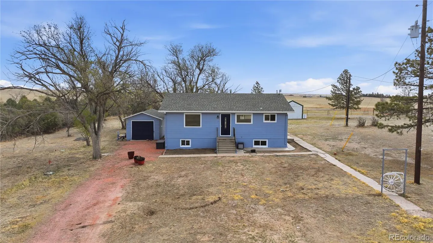 A blue single-story house with a detached garage on a large, dry rural lot under a clear blue sky.