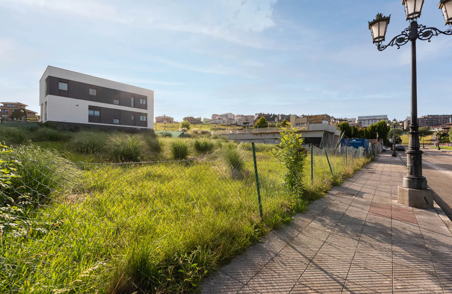 A grassy vacant lot enclosed by a chain-link fence next to a sidewalk with a street lamp, near a modern white building.