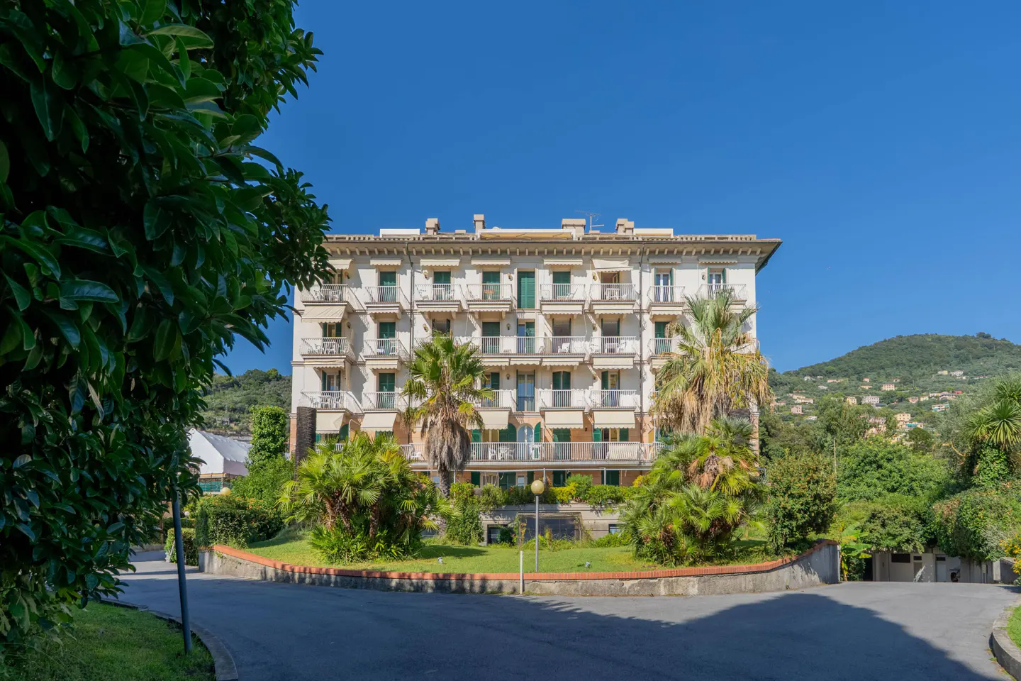 Exterior of a multi-story white building with balconies, palm trees, and a blue sky.
