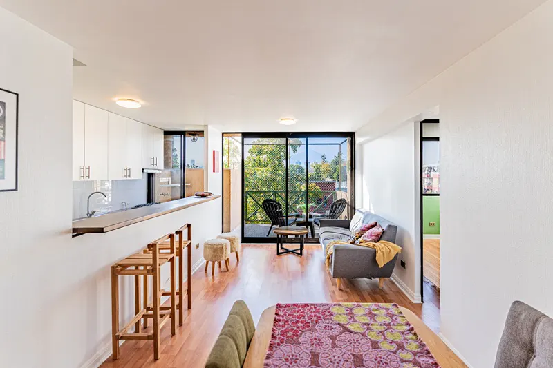 Bright apartment interior with wood floors, white walls, and a balcony with black chairs overlooking green trees. A gray sofa and wooden bar stools add warmth.