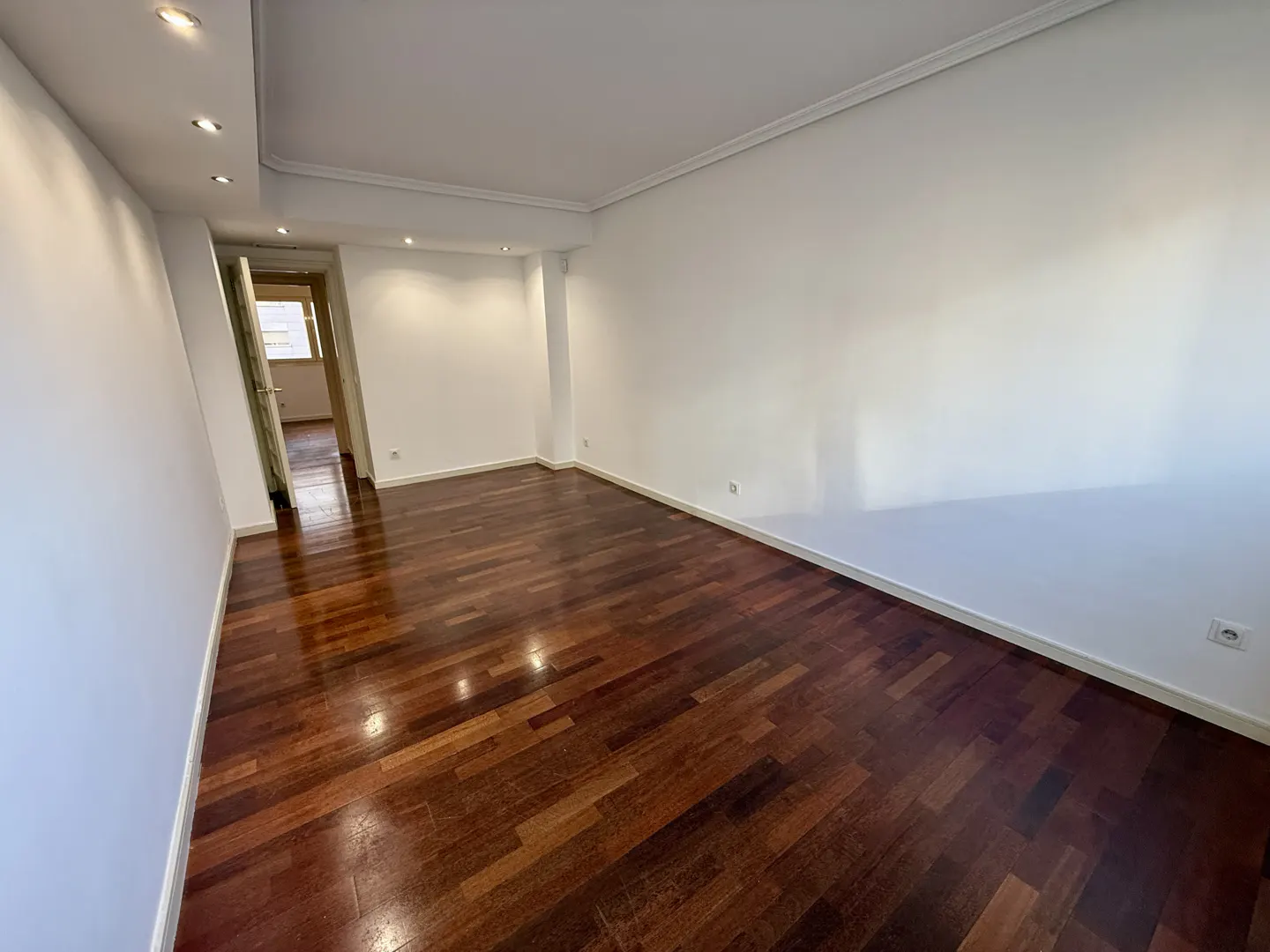 Bright, empty room with dark wood floors and white walls. A doorway leads to another room. Recessed lighting in the ceiling.