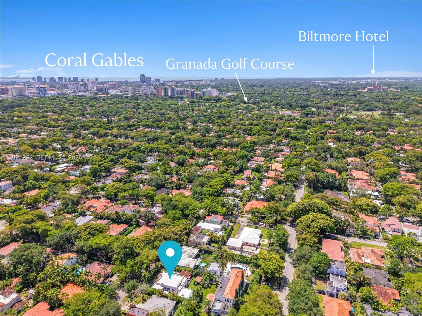 Aerial view of a residential area with a blue location pin on a white house, surrounded by green trees. Coral Gables and Biltmore Hotel are in the background.
