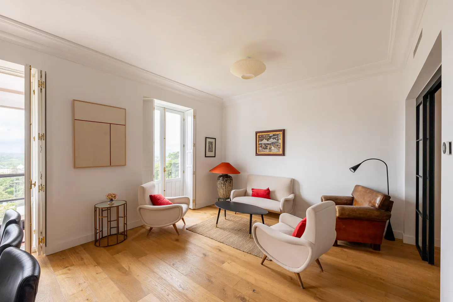 Bright living room with hardwood floors, white walls, and neutral furniture accented with red pillows. Balcony view visible through open doors.