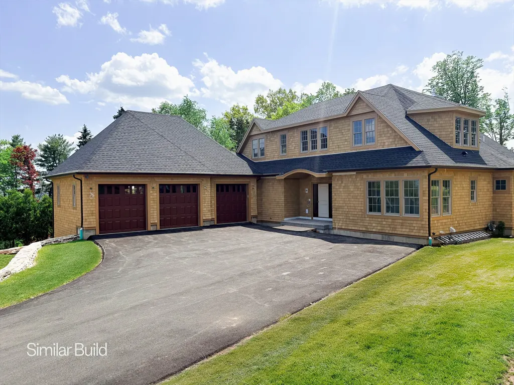 Tan shingle house with a black roof, three-car garage, and a large asphalt driveway. Green lawn and trees surround the house.