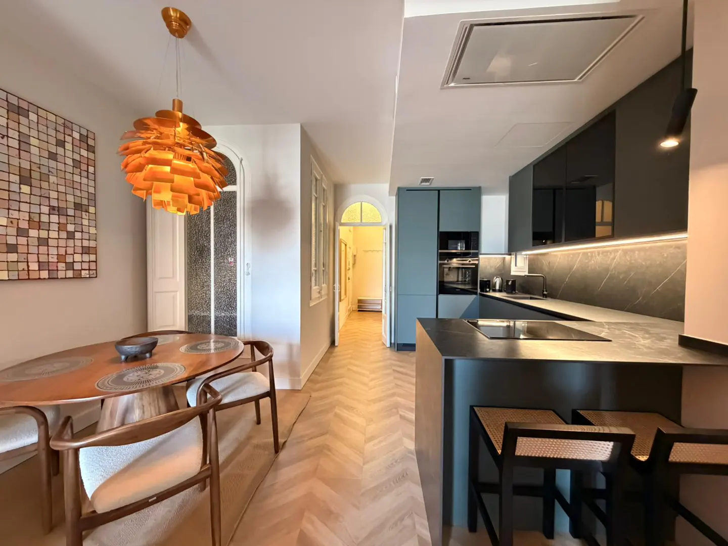 Open-concept apartment with a dining area featuring a wooden table, chairs, and a modern orange pendant light. A sleek kitchen with dark cabinets is visible.