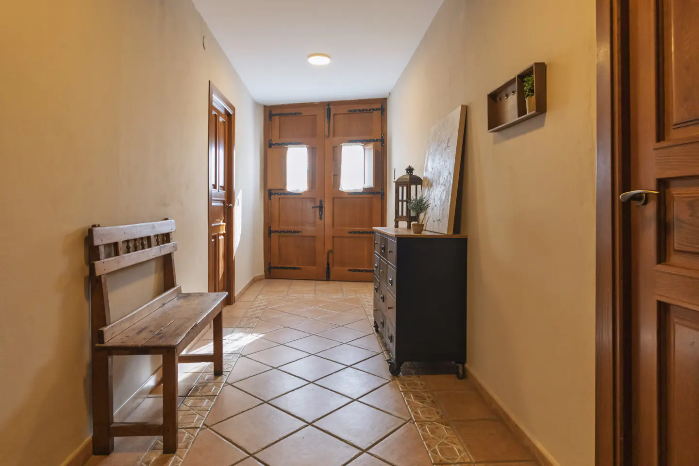 Hallway with terracotta tile floor, wooden bench, black dresser, and double wooden doors with small windows.