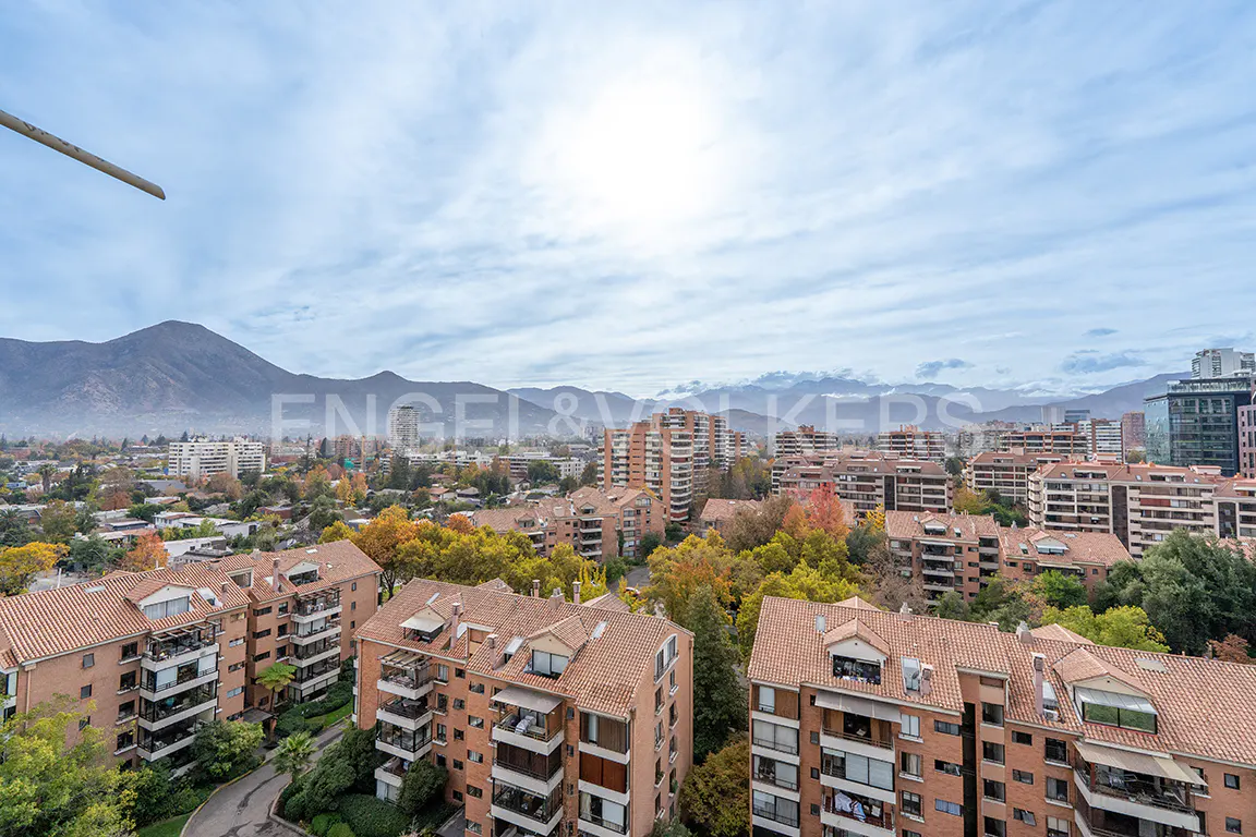 Cityscape view of Santiago, Chile, with red-roofed buildings, trees, and the Andes Mountains in the background under a cloudy sky.