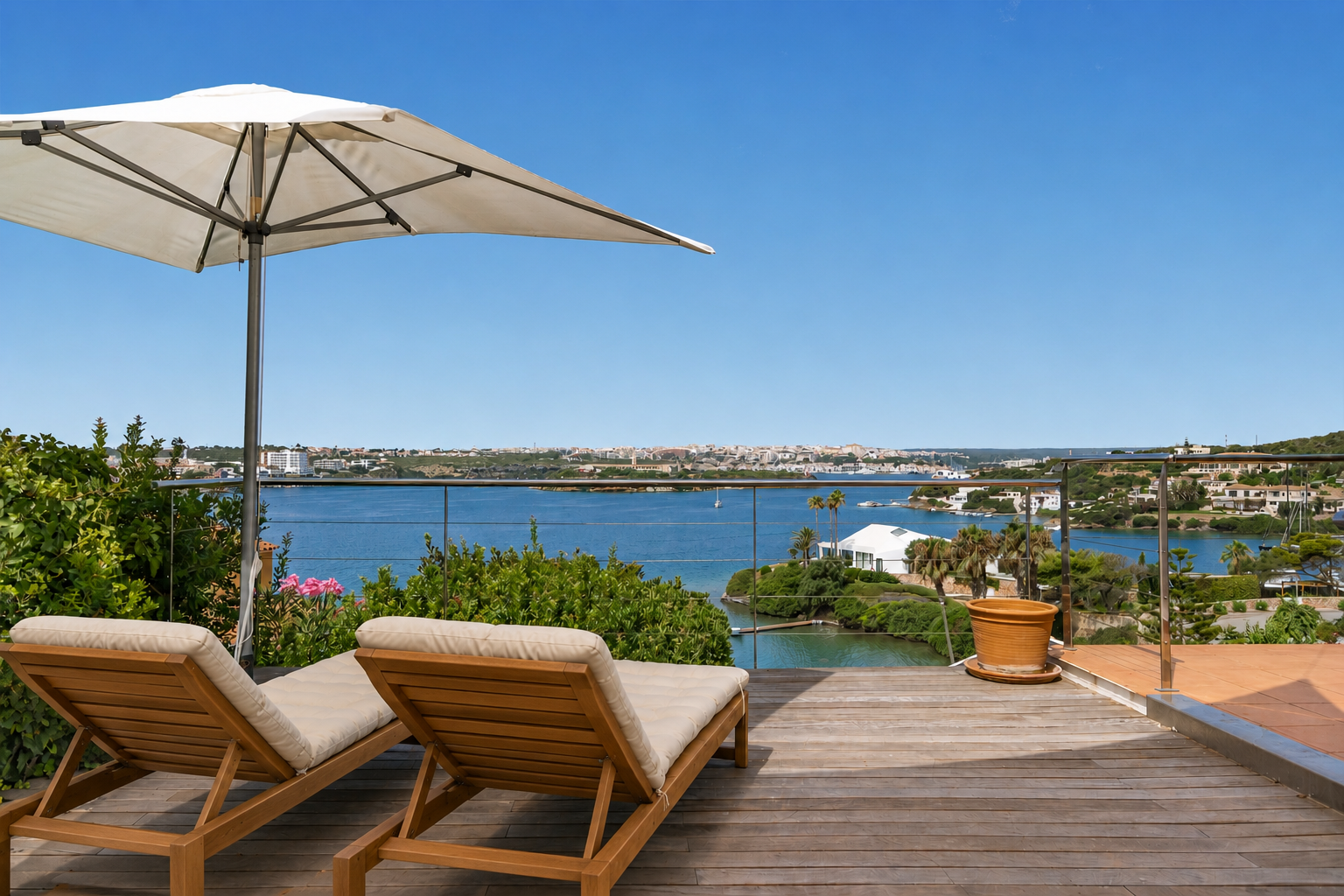 Two wooden lounge chairs with beige cushions under a white umbrella on a deck overlooking a scenic bay and distant town.