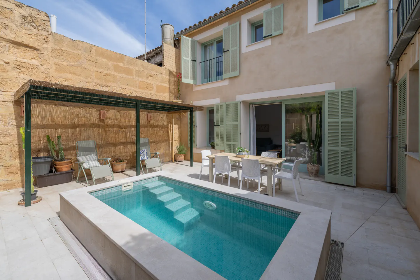 Outdoor patio with a small pool, dining table, and lounge chairs. The building has light green shutters and a balcony.