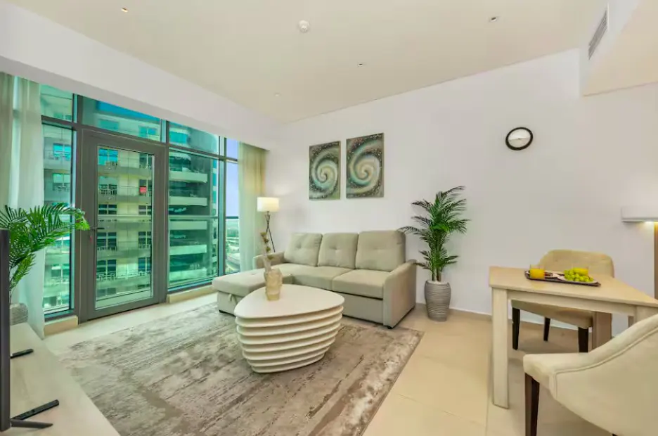 Bright apartment living room with beige sofa, unique coffee table, dining set, and city view through large windows.