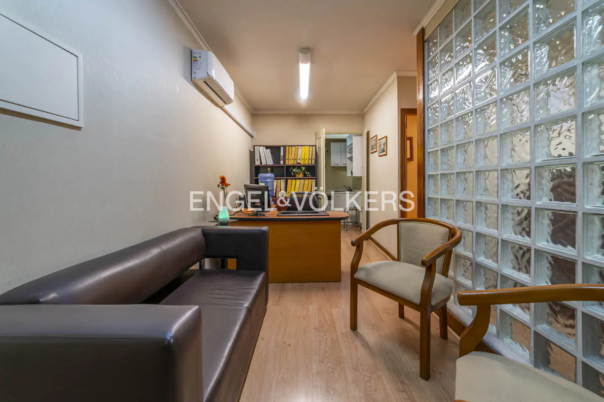 Office space with a brown leather sofa, wooden chairs, and a desk with shelves in the background. A glass block wall is on the right.