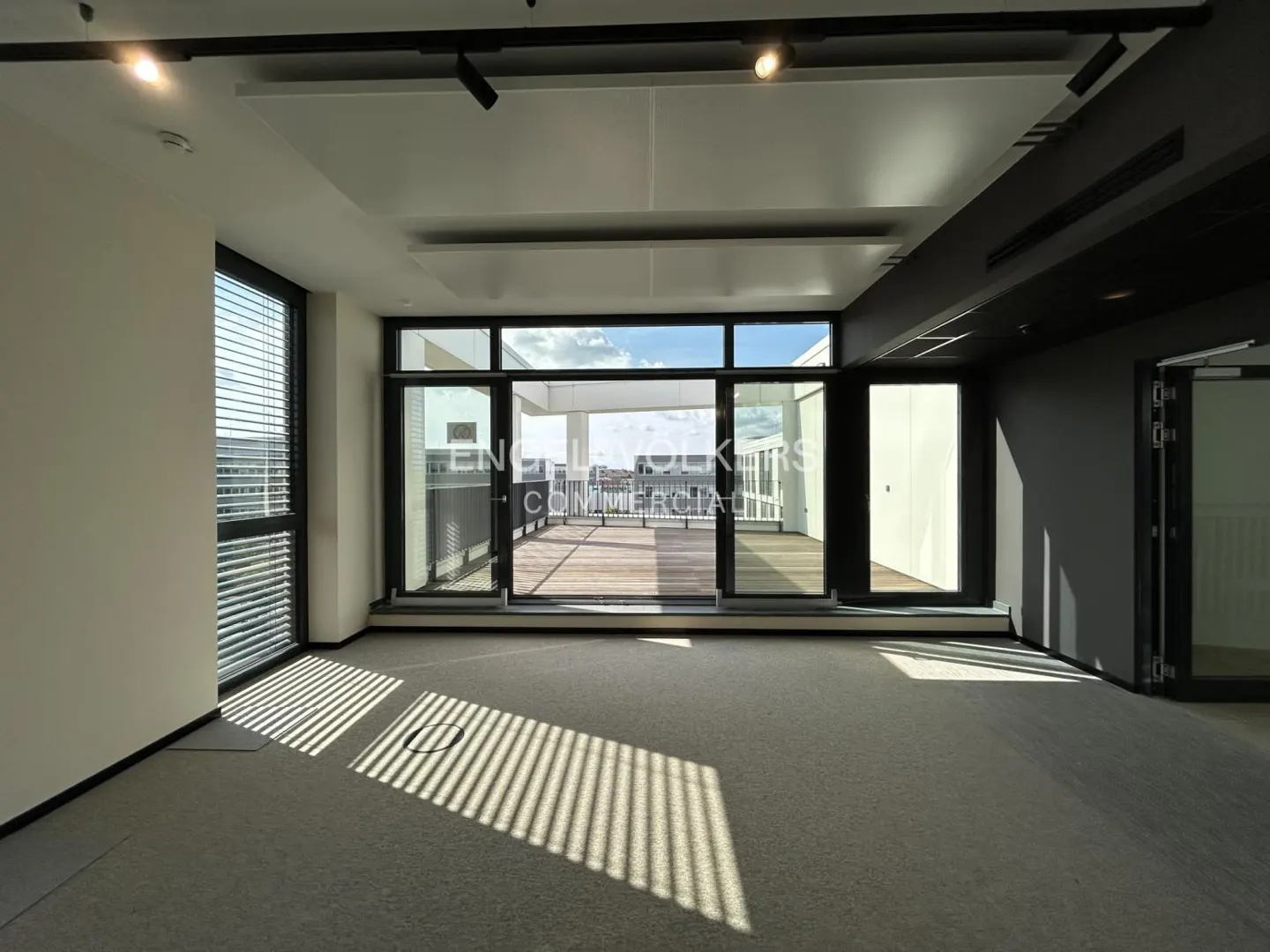 An empty office space with large windows and a balcony. Sunlight streams through blinds onto the gray carpet. The ceiling has track lighting.