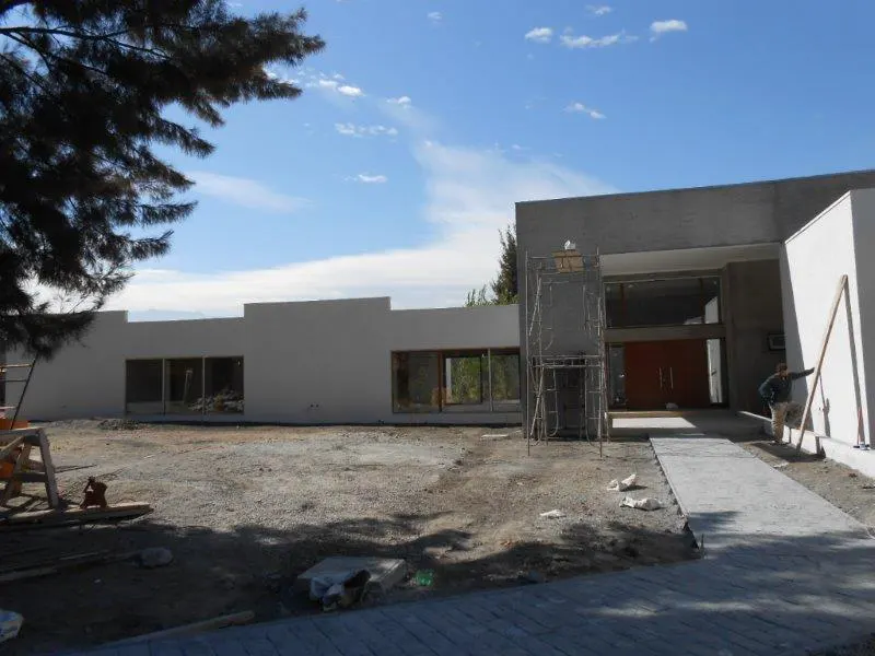 Exterior view of a modern, gray house under construction with a worker leaning against the wall. Scaffolding is visible near the entrance.