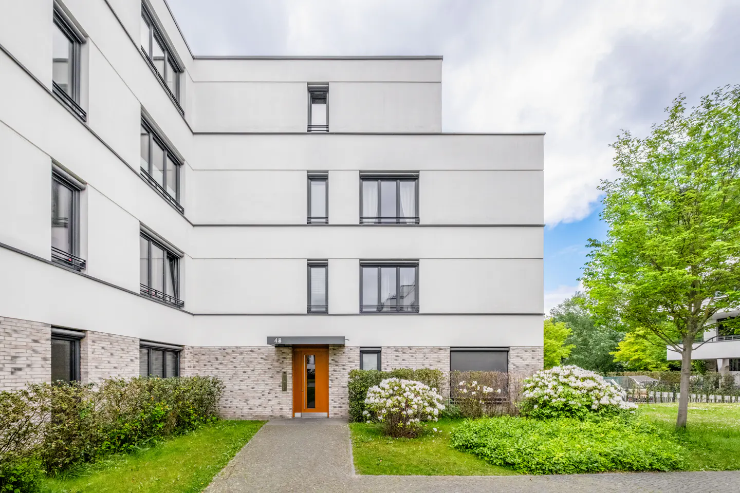 Exterior of a modern white apartment building with a brown door, gray windows, and green landscaping.