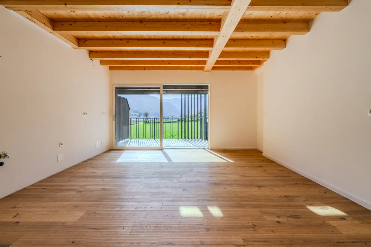 Empty room with wood floors, white walls, and exposed wood beam ceiling. Sliding glass doors lead to a balcony with a green field view.