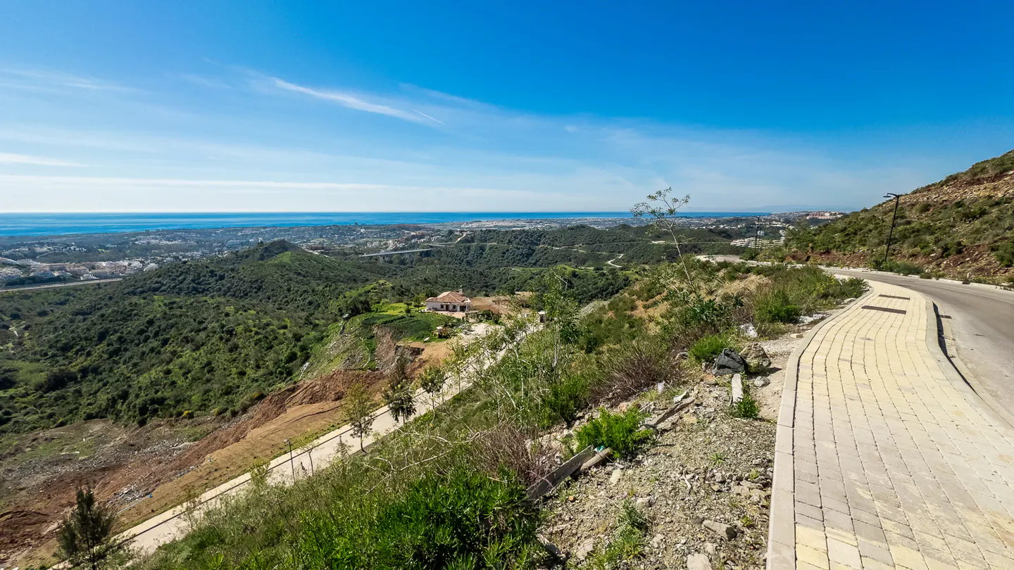 Scenic view of a hillside property with a paved road, lush greenery, and a distant ocean view under a clear blue sky.
