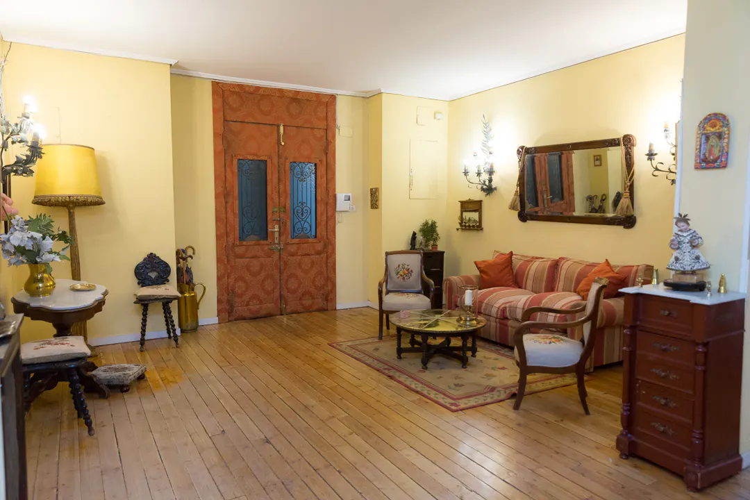 A living room with yellow walls, wooden floors, and antique furniture. A striped sofa and chairs sit on a rug. A large, patterned door is visible.