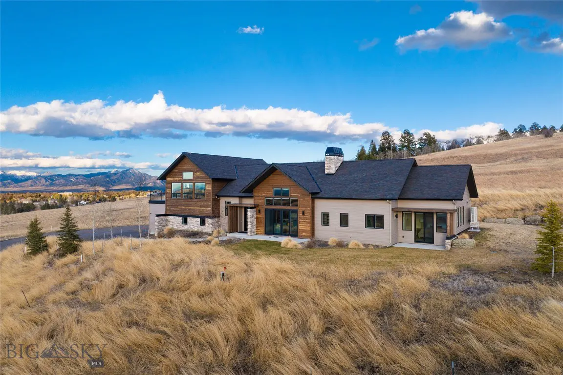 Exterior view of a modern home with wood and stone accents, set on a grassy hill under a blue sky with clouds. Mountains are visible in the distance.