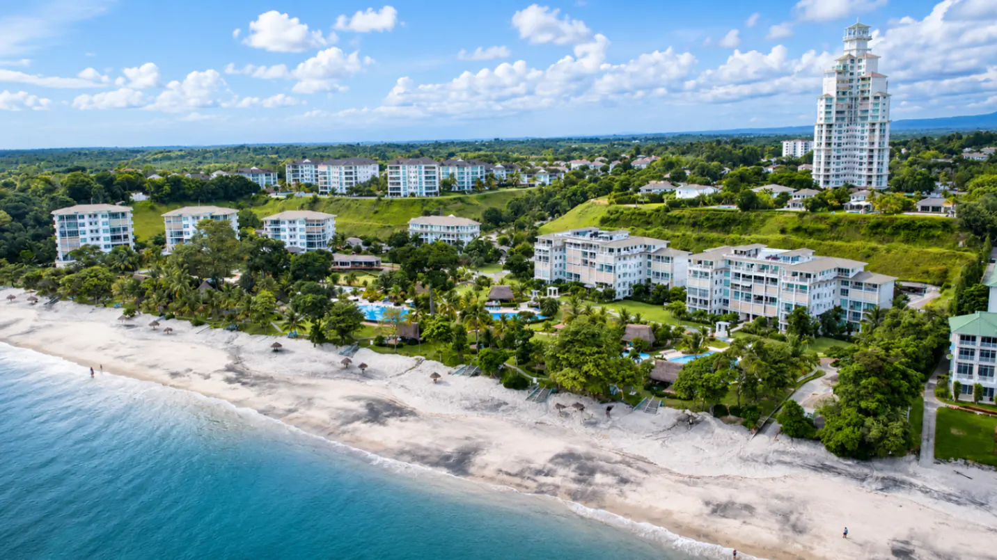Aerial view of a white sand beach, turquoise ocean, and luxury apartment complexes nestled in lush green hills.