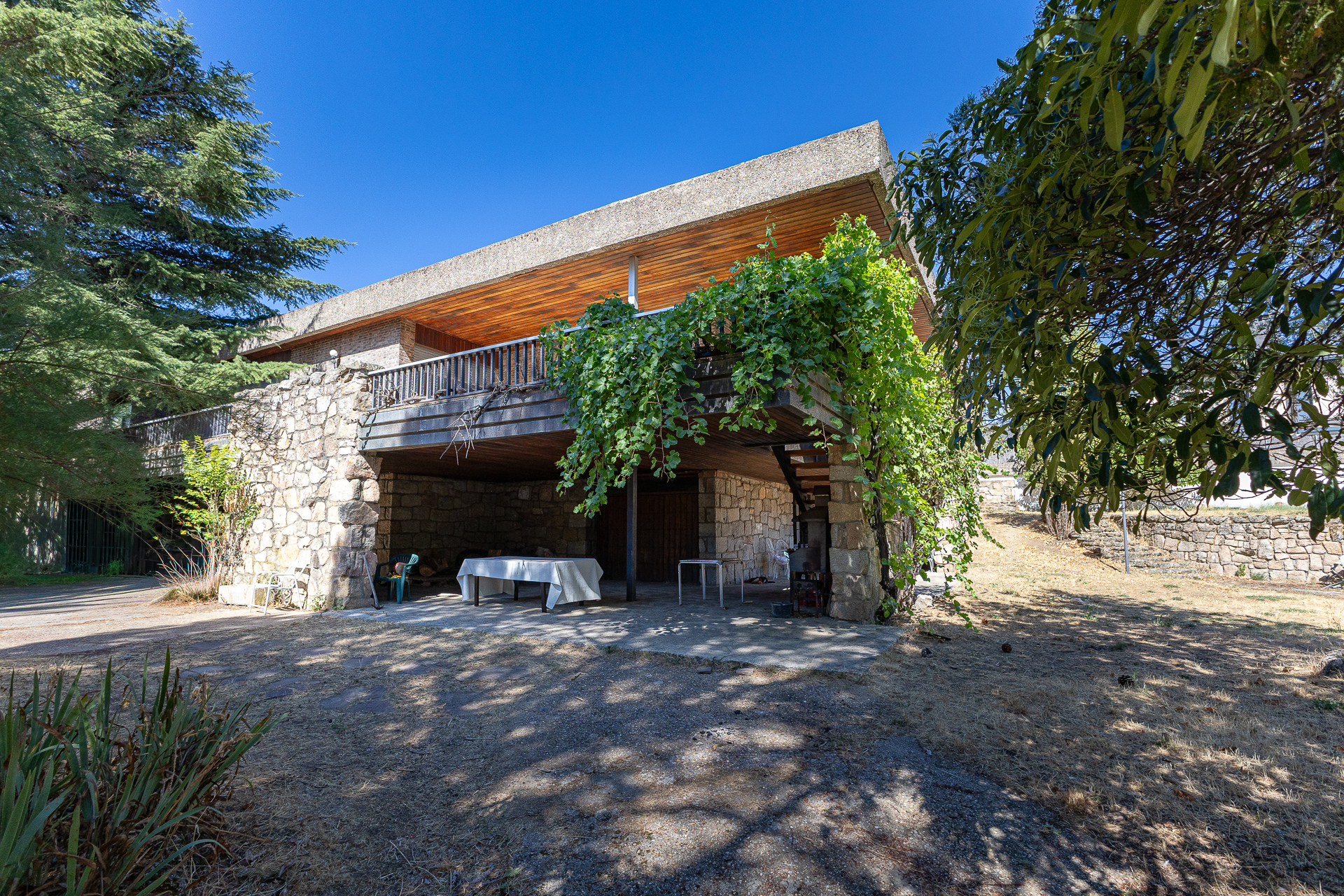 Modern stone house with a wooden ceiling balcony, covered patio, white table, and lush greenery under a clear blue sky.
