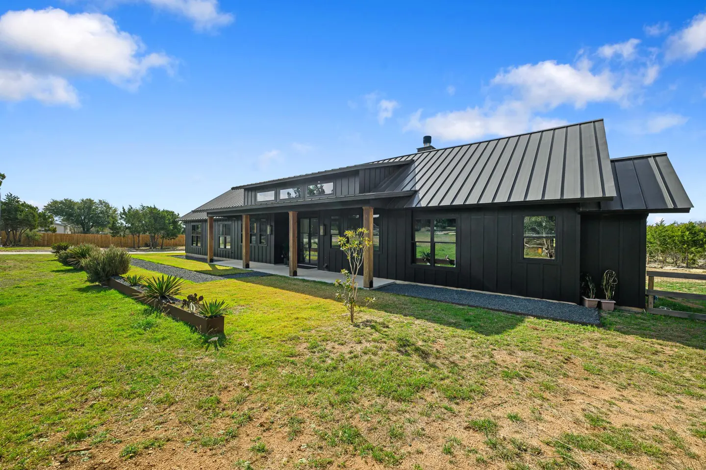 Modern black farmhouse with a metal roof, wooden porch pillars, and a grassy yard under a bright blue sky.
