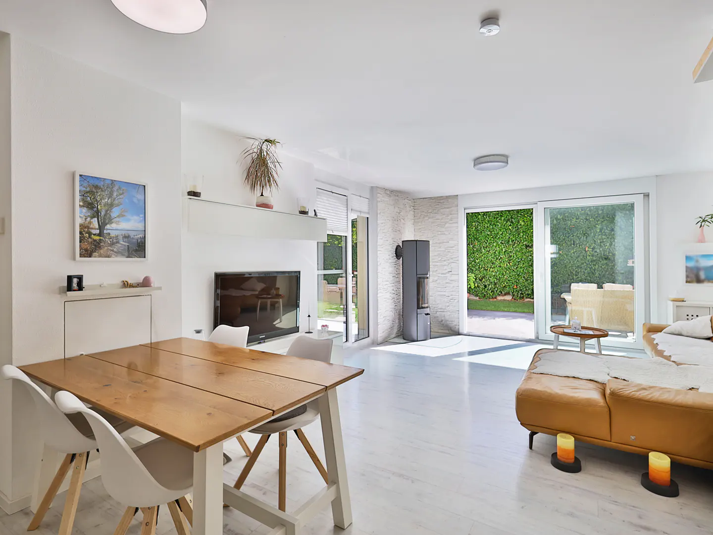 Bright, open-plan living area with a wooden dining table, white chairs, and a tan leather sofa. Sliding glass doors lead to a green garden.