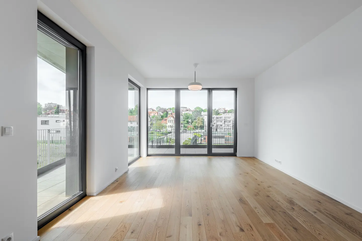 Bright, empty room with light wood floors, white walls, and large black-framed windows overlooking a cityscape. A modern light fixture hangs from the ceiling.