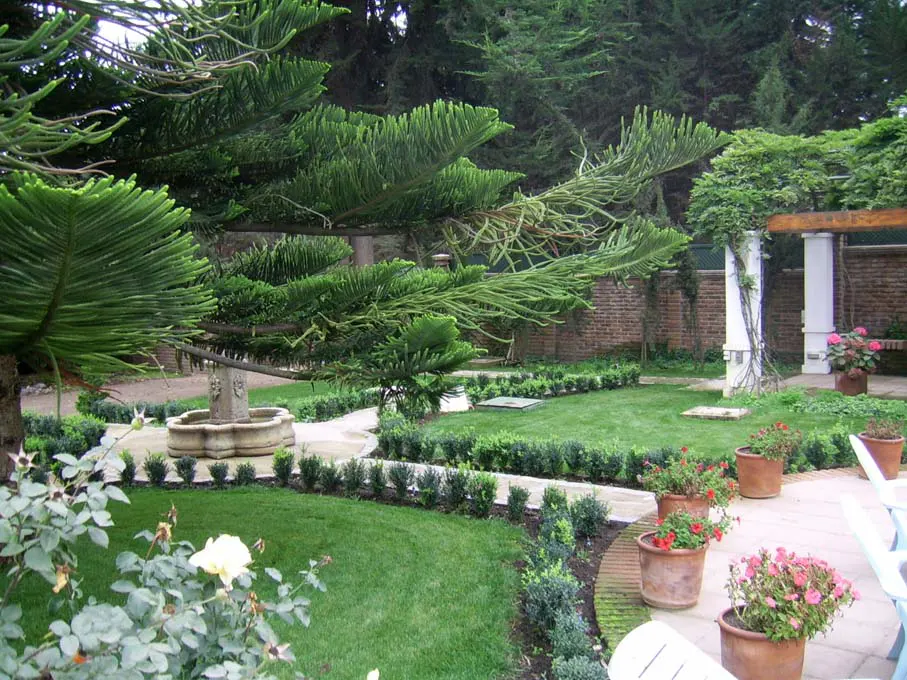 A lush green garden with a stone fountain, manicured hedges, and potted flowers. A pergola stands in the background.
