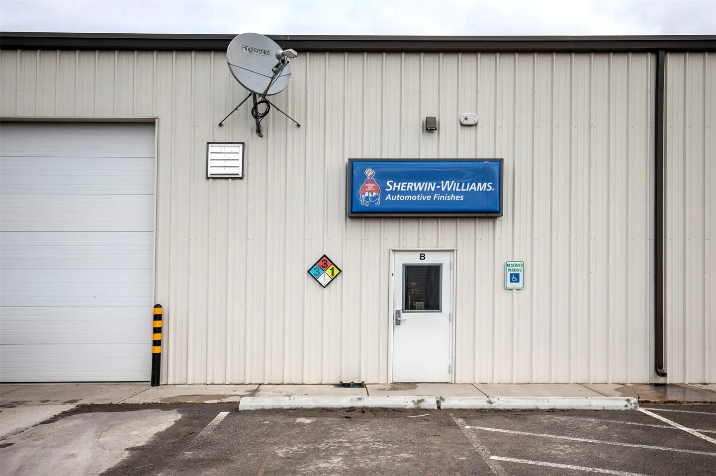 Exterior of a Sherwin-Williams Automotive Finishes building with a white metal facade, a bay door, and a parking lot.