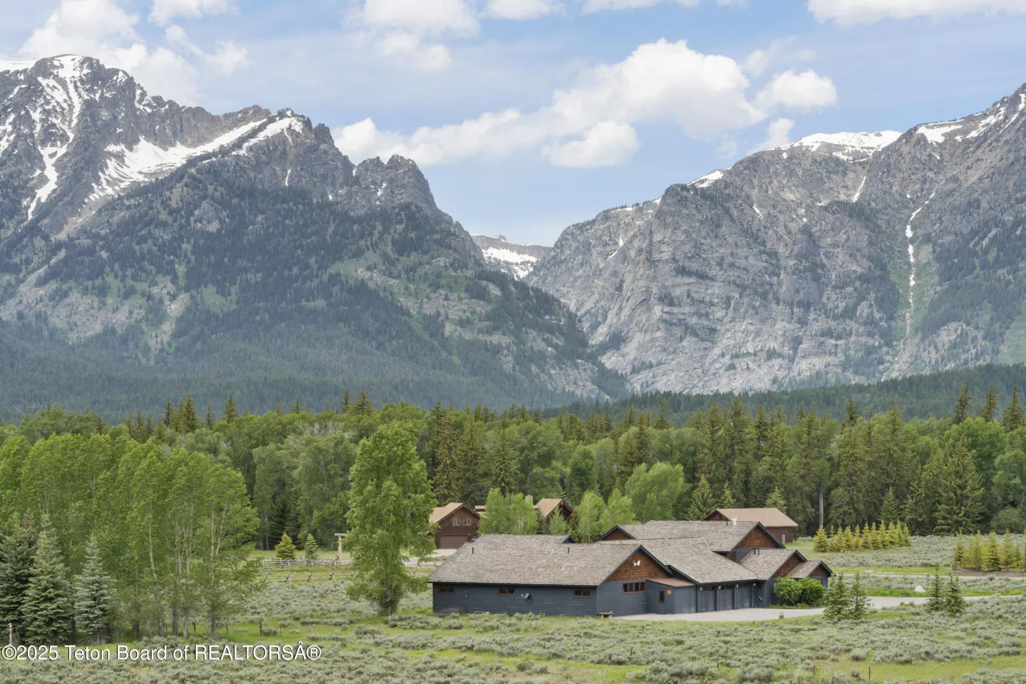 A gray house sits in a field with mountains and trees in the background.