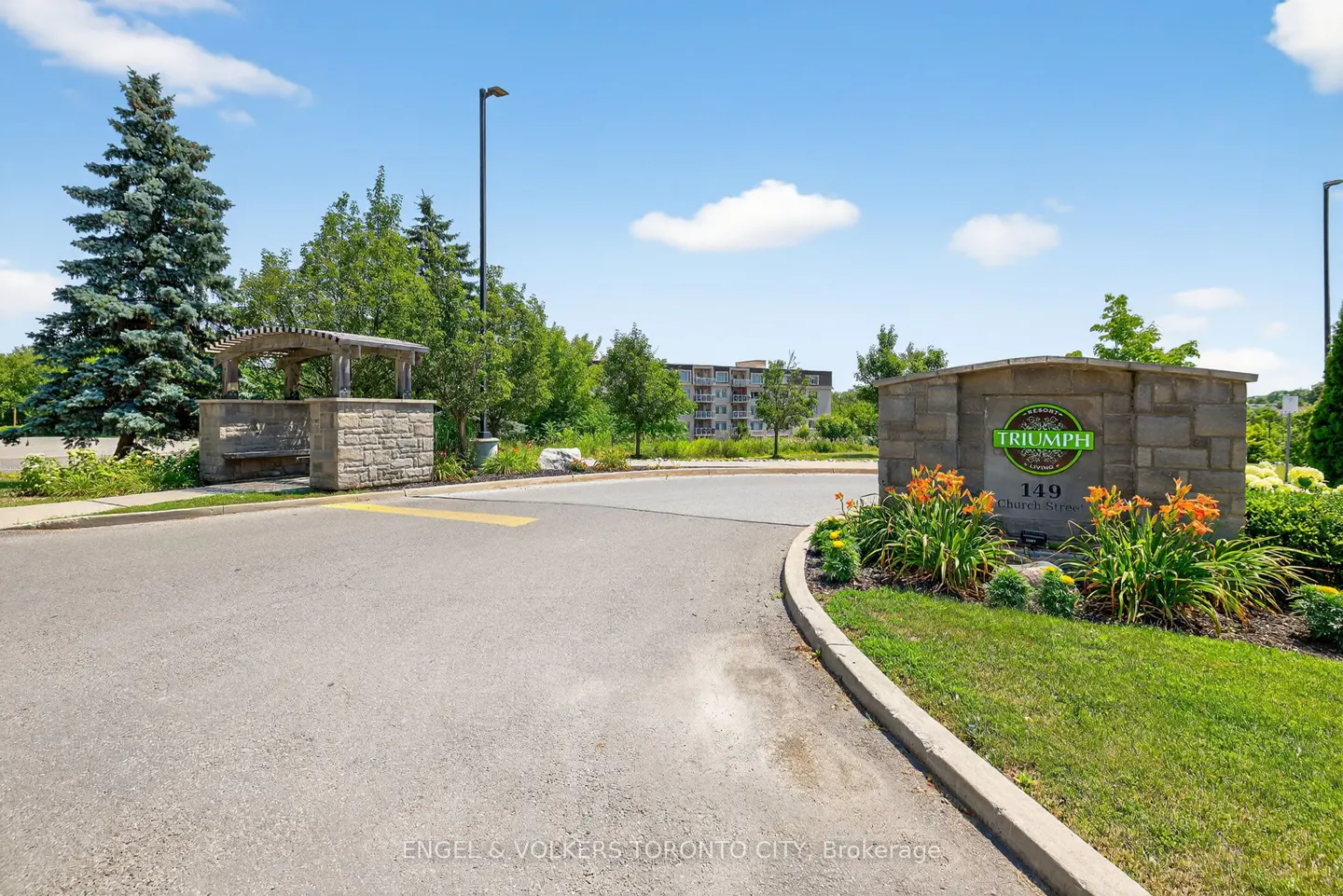 Stone entrance sign for Triumph at 149 Church Street with a paved driveway, landscaping, and a wooden pergola.
