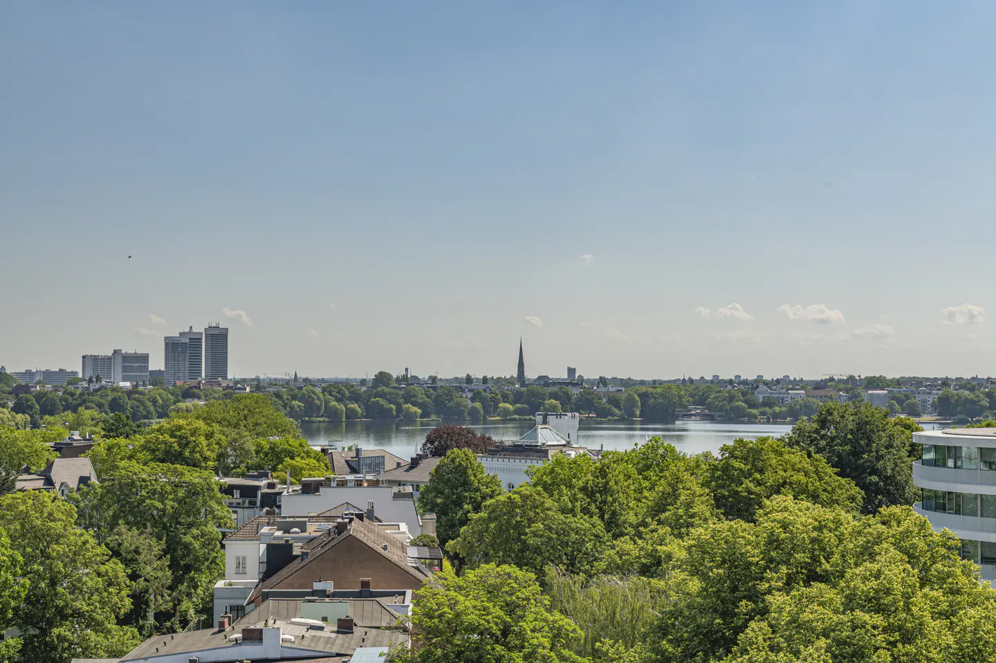 View of Hamburg, Germany, with rooftops and green trees in the foreground, a lake in the middle ground, and city buildings in the background under a blue sky.
